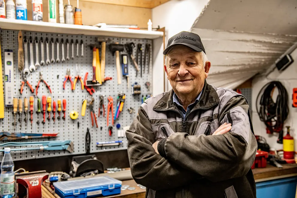 Älterer Mitarbeiter in Arbeitskleidung steht mit verschränkten Armen in einer Werkstatt vor einer Werkzeugwand mit Zangen, Schraubenschlüsseln und Hämmern