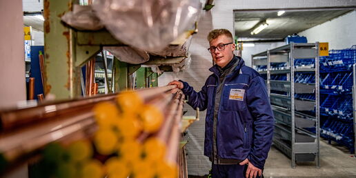 Mitarbeiter in blauer Arbeitskleidung steht im Lager neben Kupferrohren und Regalen mit blauen Kunststoffboxen.
