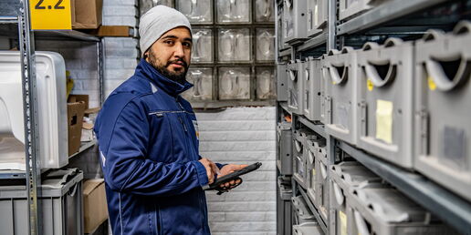 Mitarbeiter in blauer Arbeitskleidung und Mütze kontrolliert mit einem Tablet Lagerbehälter in einem Regallager