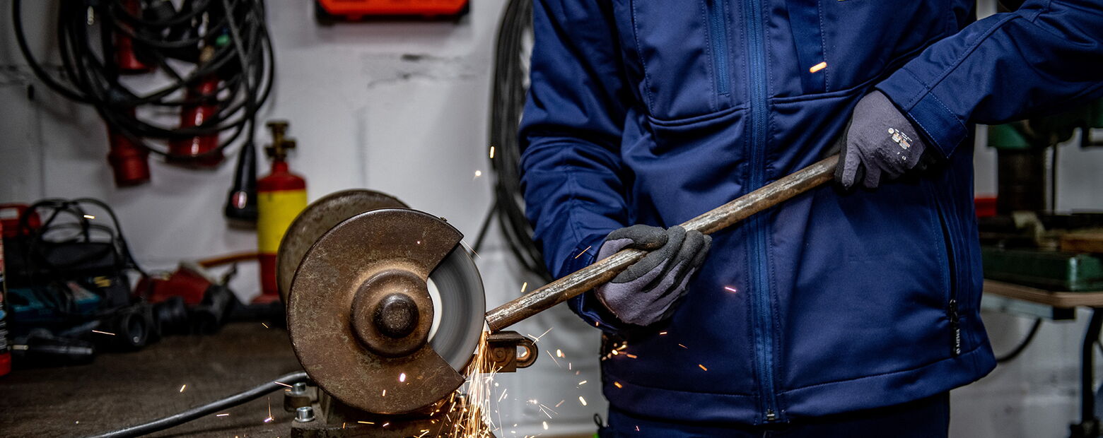 Mitarbeiter in blauer Arbeitskleidung schleift ein Metallrohr an einer Trennschleifmaschine, Funken fliegen beim Schleifen.