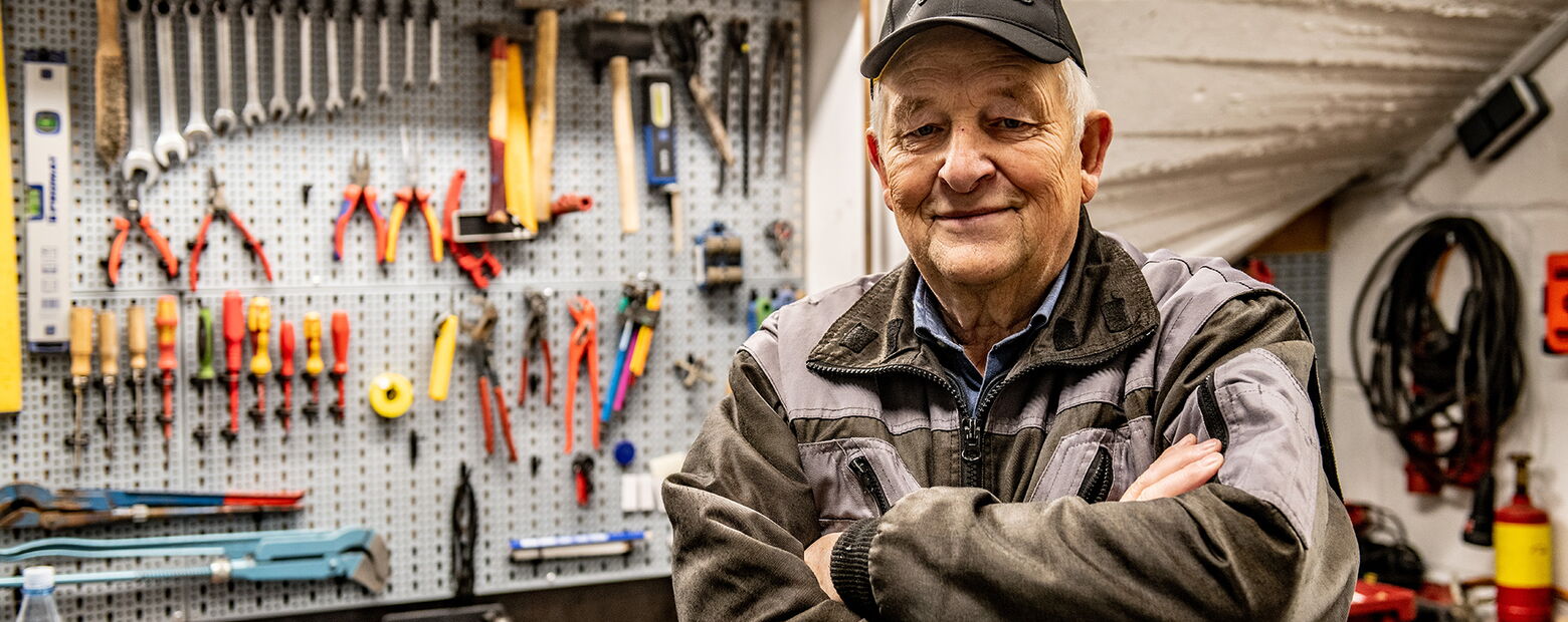 Älterer Mitarbeiter in Arbeitskleidung steht mit verschränkten Armen in einer Werkstatt vor einer Werkzeugwand mit Zangen, Schraubenschlüsseln und Hämmern